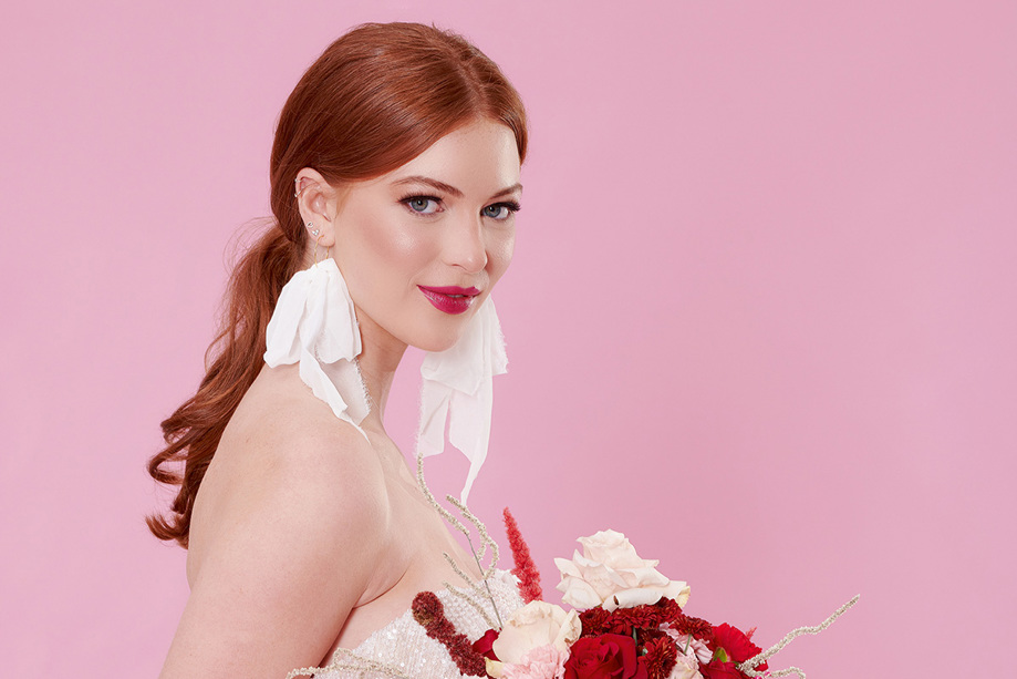 Model poses with bouquet in front of pink backdrop wearing fabric draped earrings